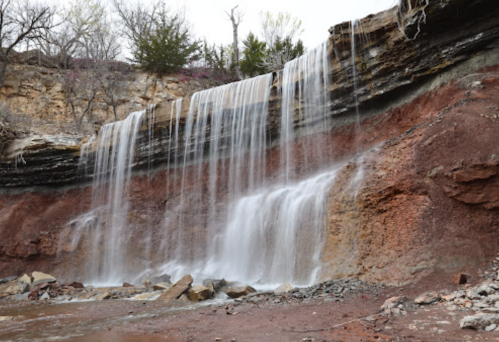 A cascading waterfall flows over rocky cliffs, surrounded by sparse trees and earthy tones.