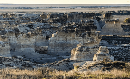 A rugged landscape of eroded rock formations and cliffs under a clear sky, with grassy plains in the background.