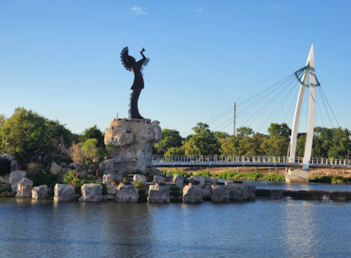 A silhouette statue on a rock by a river, with a modern bridge in the background under a clear blue sky.