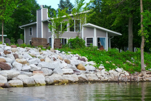 A lakeside house surrounded by greenery and large rocks, with a calm water view in the foreground.