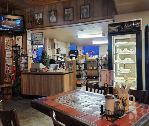 Cozy diner interior with a tiled table, dessert display, and a counter, featuring rustic decor and a TV in the background.