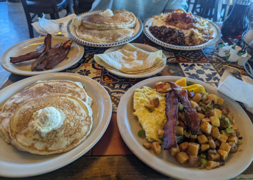 A table with various breakfast dishes: pancakes, bacon, eggs, potatoes, and tortillas, all served on colorful plates.
