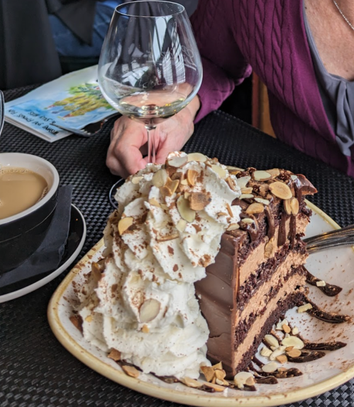 A slice of chocolate cake topped with whipped cream and almonds, alongside a cup of coffee and a wine glass.