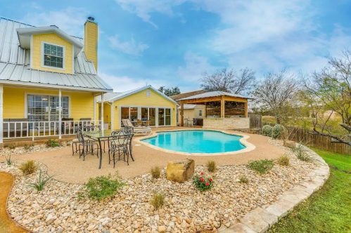A sunny backyard with a pool, surrounded by rocks and plants, featuring a yellow house and a covered patio area.