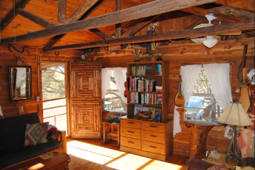 Cozy wooden interior of a cabin with a bookshelf, rustic furniture, and large windows letting in natural light.