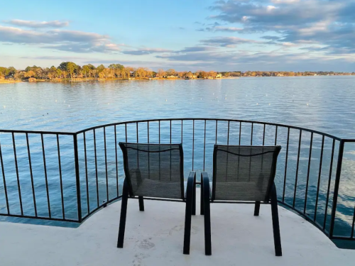 Two chairs on a balcony overlooking a calm lake with trees and clouds in the background.
