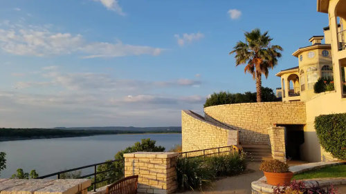 Scenic view of a lake with a stone pathway, palm tree, and a house against a blue sky with scattered clouds.