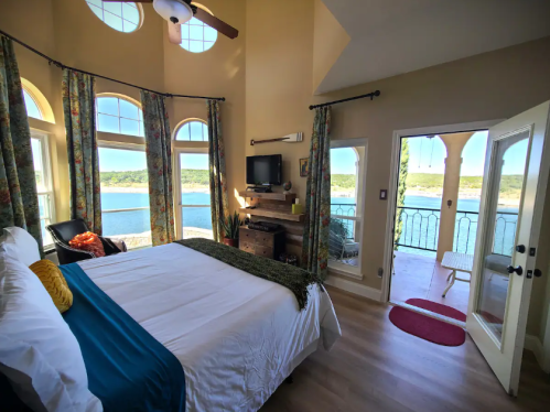 Cozy bedroom with large windows overlooking a lake, featuring a bed, chair, and balcony access. Bright and inviting space.