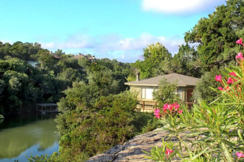 A serene landscape featuring a house by a lake, surrounded by lush greenery and blooming flowers under a blue sky.