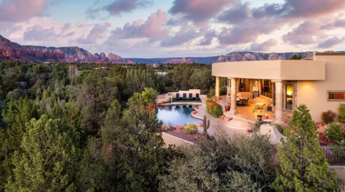 A scenic view of a modern home surrounded by trees, with a pool and mountains in the background under a colorful sky.