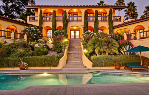 A beautiful villa with a landscaped garden, stairs leading to a grand entrance, and a pool in the foreground at dusk.