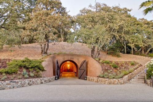 A rustic entrance with large wooden doors set into a hillside, surrounded by trees and a stone pathway.