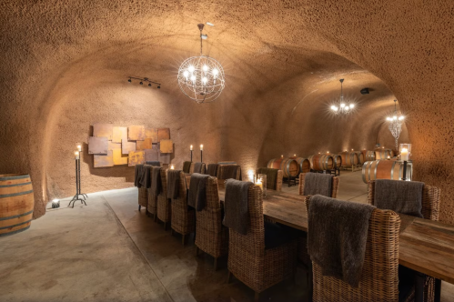 A rustic wine cellar with a long table, wicker chairs, and wooden barrels, illuminated by stylish chandeliers.