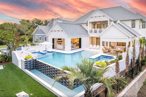 A modern home with a large pool, surrounded by palm trees and a well-manicured lawn, under a colorful sunset sky.