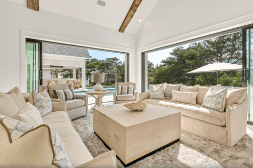 Bright, modern living room with beige sofas, a wooden coffee table, and large windows overlooking greenery.
