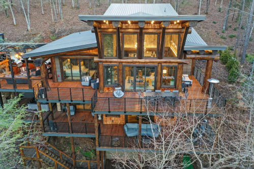 A modern wooden cabin with large windows, multiple decks, and a forest backdrop.