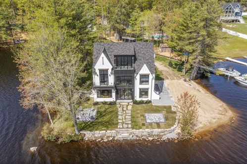 A modern house by a lake, surrounded by trees, with a stone pathway and outdoor seating area.