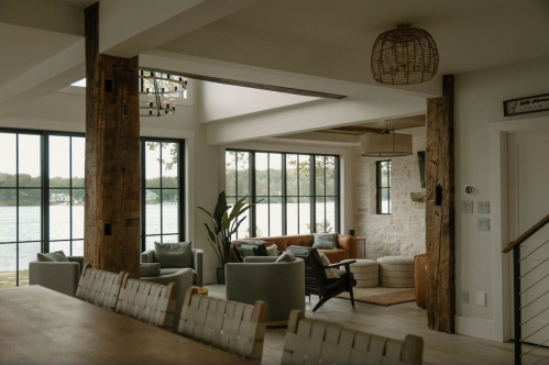 A modern living space with large windows, wooden beams, and cozy seating overlooking a lake.