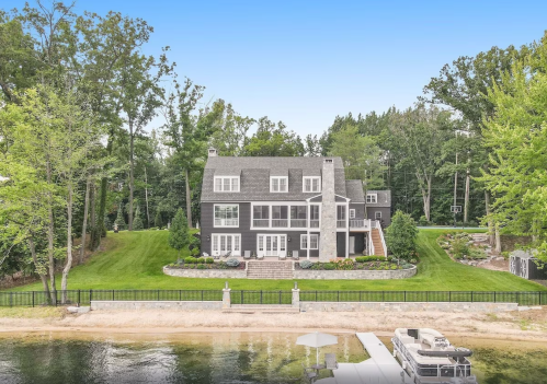 A large, modern black house surrounded by trees, with a lawn leading to a sandy beach and water in the foreground.