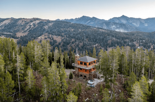 A wooden cabin surrounded by tall trees, set against a backdrop of mountains and a clear sky.