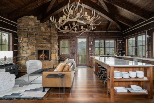 Cozy rustic kitchen and living area with stone fireplace, wooden beams, and large windows showcasing mountain views.