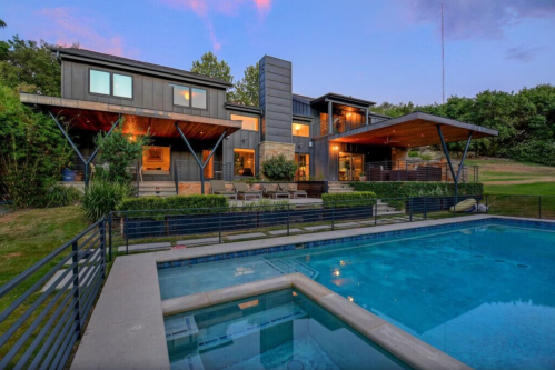 Modern house with large windows, surrounded by greenery, featuring a pool in the foreground at dusk.
