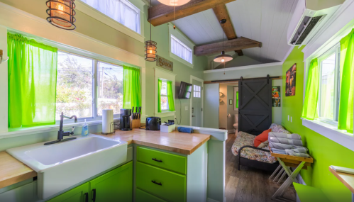 Bright kitchen with green walls, wooden beams, and a cozy living area visible through an open doorway.