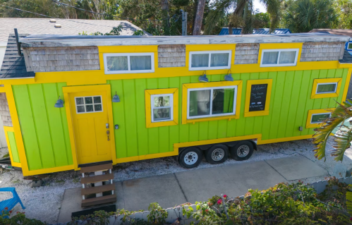A vibrant green and yellow tiny house on wheels, surrounded by palm trees and a gravel path.