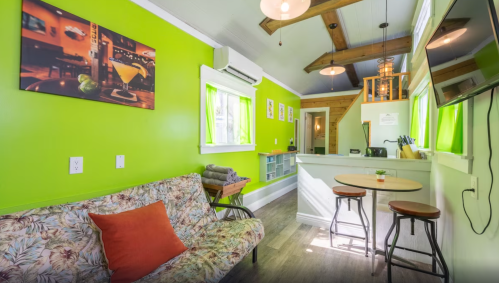 Cozy green living space with a floral couch, bar stools, and a kitchenette, featuring wooden beams and bright decor.