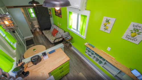 A bright green room with a cozy seating area, wooden table, and kitchen space, featuring large windows and decorative art.