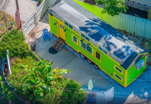 A vibrant green and yellow tiny house on wheels, surrounded by greenery and a concrete patio.