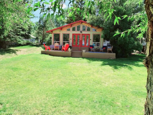 A charming cabin with red doors and a deck, surrounded by green grass and trees on a sunny day.