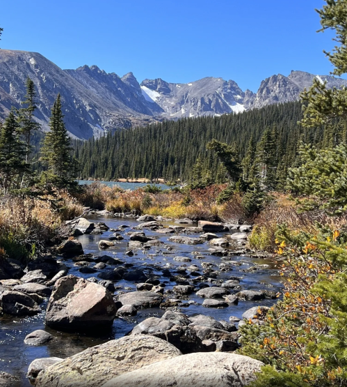 A serene mountain landscape with a clear stream, rocky banks, and towering peaks under a bright blue sky.