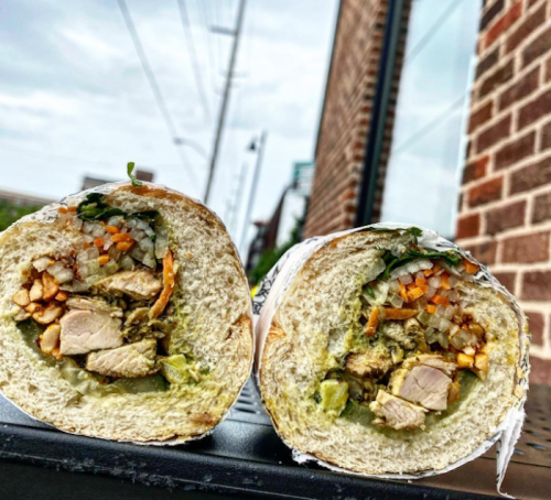 A close-up of a halved sandwich filled with grilled chicken, vegetables, and herbs, resting on a table outdoors.