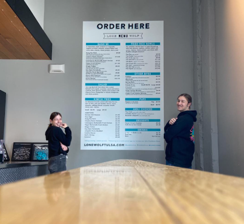 Two people stand beside a large menu board in a restaurant, with a wooden table in the foreground.