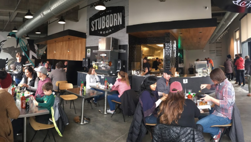 A busy restaurant interior with diners at tables, a soda fountain, and a kitchen area in the background.