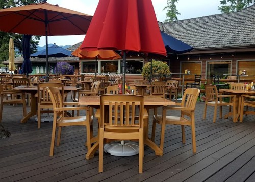 Outdoor dining area with wooden tables and chairs, shaded by large umbrellas, surrounded by trees.