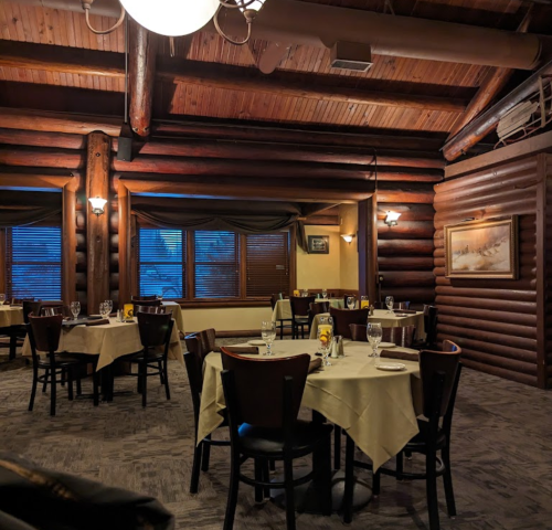 Cozy restaurant interior with wooden log walls, tables set with white linens, and warm lighting.