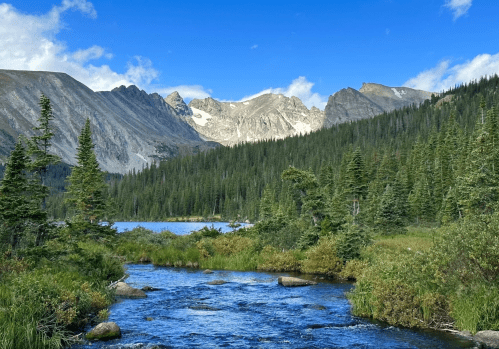 A serene mountain landscape with a clear stream, lush greenery, and towering peaks under a blue sky with clouds.