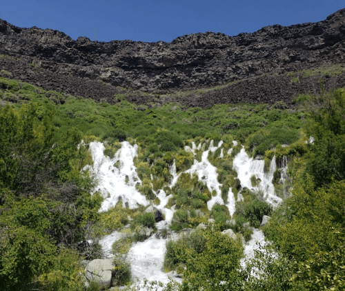 A cascading waterfall flows down a rocky cliff, surrounded by lush greenery under a clear blue sky.