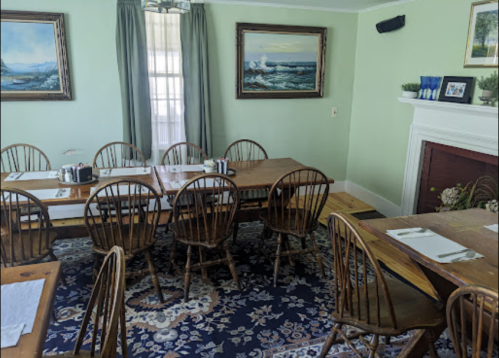 A cozy dining room with wooden tables and chairs, green walls, and paintings of seascapes.