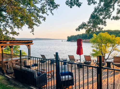 A serene lakeside view with a wooden deck, lounge chairs, and a red umbrella under a clear sky.