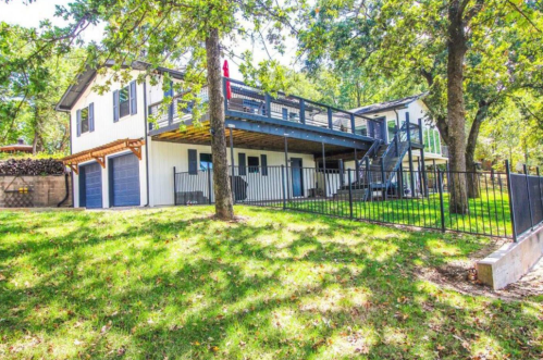 A two-story house with a deck, surrounded by trees and a grassy yard, featuring a fenced area and two garage doors.
