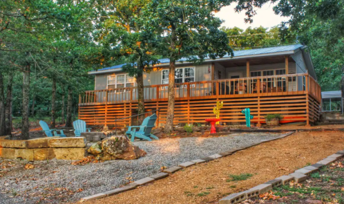 A cozy cabin with a wooden deck, surrounded by trees and colorful outdoor furniture, set on a gravel path.