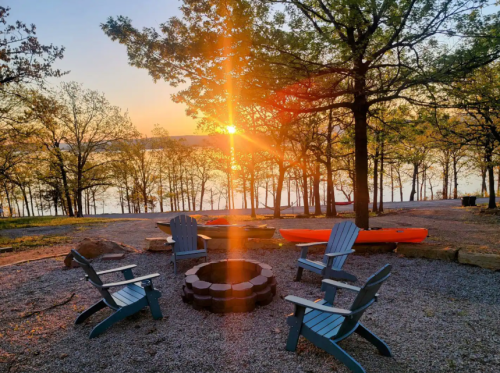 A serene lakeside scene at sunset, featuring a fire pit surrounded by blue chairs and a kayak nearby.