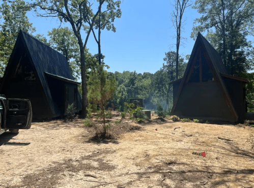 Two A-frame cabins surrounded by trees on a sunny day, with a clearing and a small fire pit in the background.