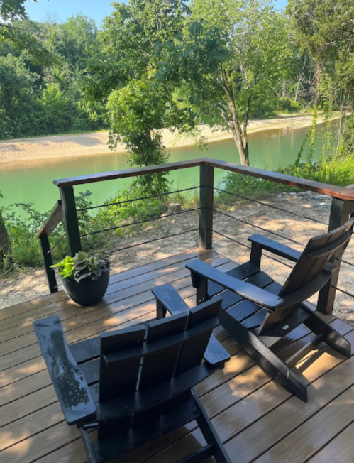 Two black Adirondack chairs on a wooden deck overlooking a serene river and lush greenery.