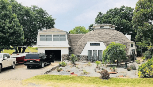A unique dome-shaped house with a stone facade, surrounded by landscaping and a driveway with a parked black SUV.