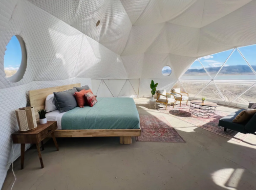 A modern, airy bedroom with a large bed, cozy seating, and panoramic views through a geodesic dome structure.