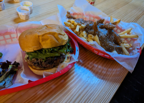 A burger with lettuce in a red basket next to a basket of fries topped with gravy and cheese curds on a wooden table.